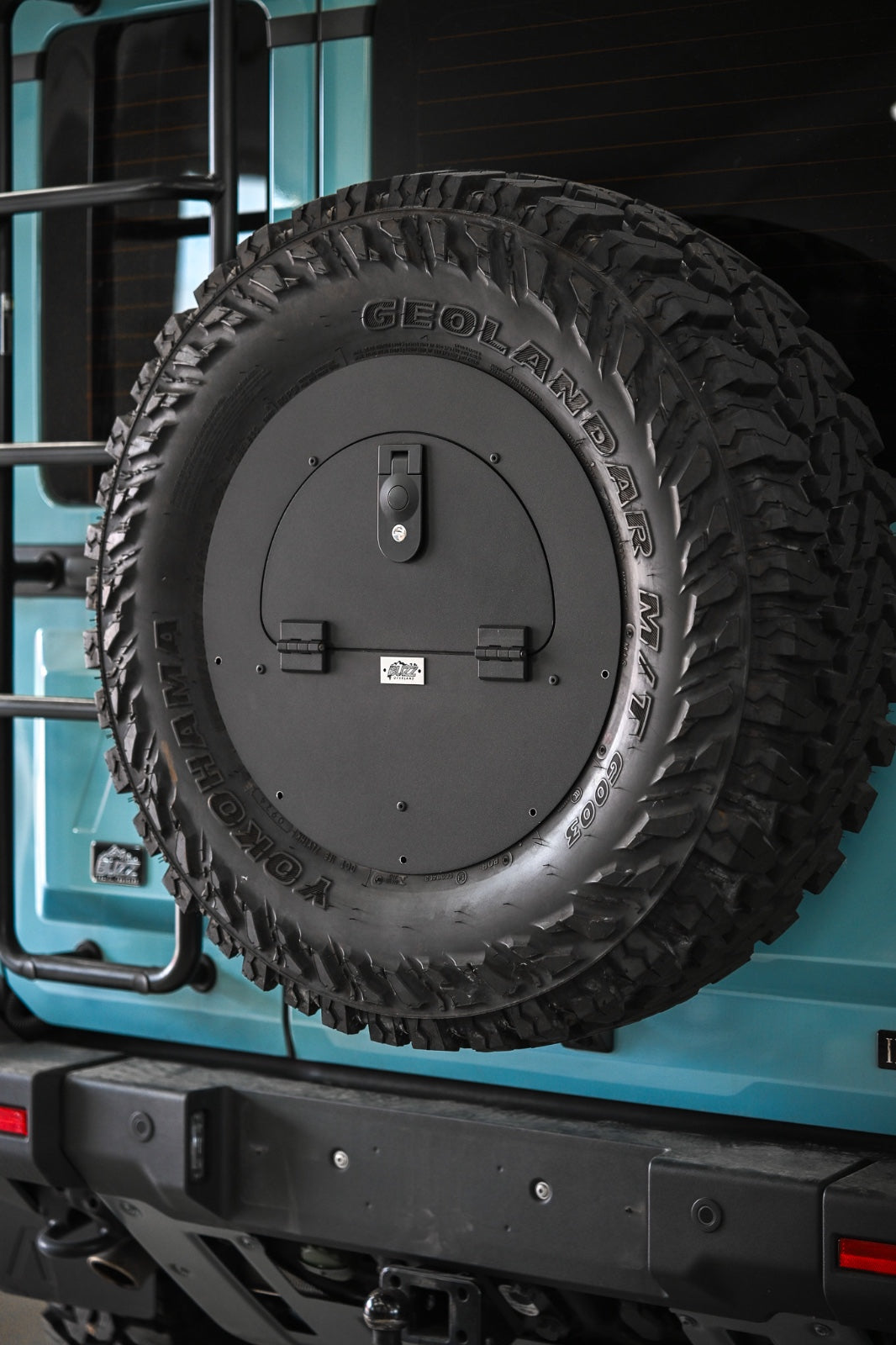 Buzz Overland Spare Wheel Storage Cubby for INEOS Grenadier, made from corrosion-resistant 5052 aluminium with a textured black powder coat, bolted to a 17-inch wheel for secure off-road storage.